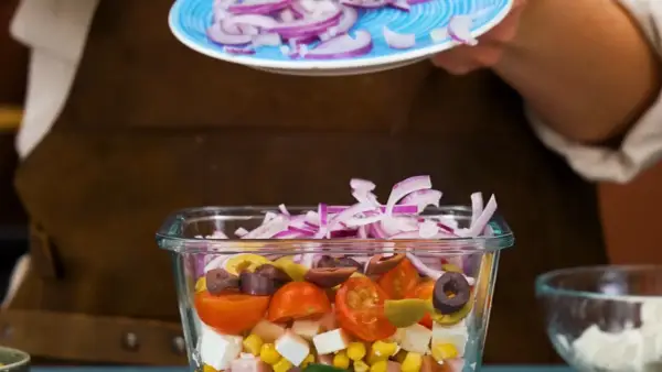 En person lägger rödlök på en pastasallad med kassler, majs, tomater, oliver och fetaost.